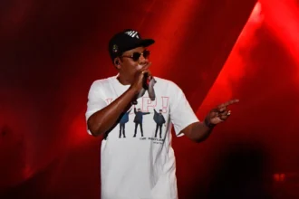 Jay-Z tritt beim Super Bowl LXI auf der Bühne auf und trägt eine Sonnenbrille, eine schwarze Baseballkappe sowie ein weißes T-Shirt mit einem Beatles-Motiv.