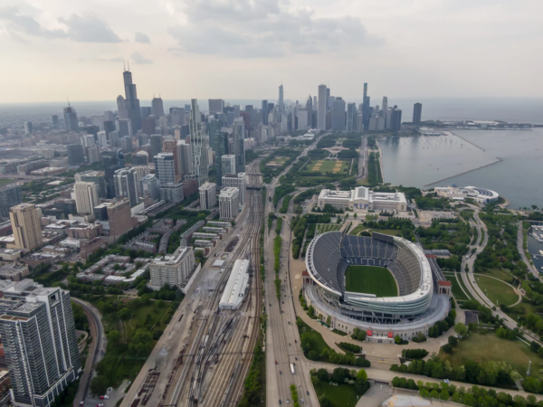 FootballR - NFL - Luftaufnahme von Chicago mit der Skyline mit hoch aufragenden Wolkenkratzern, dem Michigansee, Soldier Field, der Heimat der Chicago Bears, und mehreren zusammenlaufenden Bahngleisen an einem bewölkten Tag. Diese Beschreibung wurde automatisch generiert.