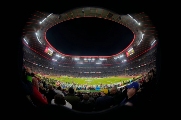 FootballR - NFL - Ein voll besetztes Footballstadion ist unter dem Abendhimmel beleuchtet. Das Spielfeld ist bereit für ein Spiel, Spieler und Offizielle sind versammelt. Fans füllen die Tribünen, viele halten Tickets für die NFL München 2024 in der Hand, während sie dem Ereignis entgegenfiebern. Der runde Rand des Stadions ist mit roten Lichtern hervorgehoben, was die elektrisierende Atmosphäre noch verstärkt. Diese Beschreibung wurde automatisch generiert.