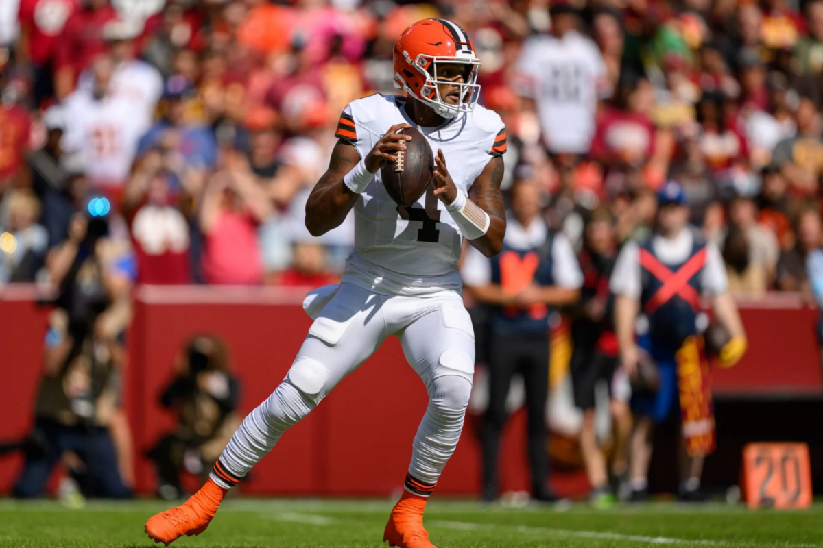 FootballR - Todd Monken - Deshaun Watson in weißer Uniform mit orangefarbenen und braunen Akzenten hält einen Football und bereitet sich zum Wurf vor. Er steht auf einem grünen Feld mit einer verschwommenen Menschenmenge im Hintergrund. Er trägt einen orangefarbenen Helm und weiße Stollenschuhe, was darauf hindeutet, dass gerade ein Spiel läuft. Diese Beschreibung wurde automatisch generiert.