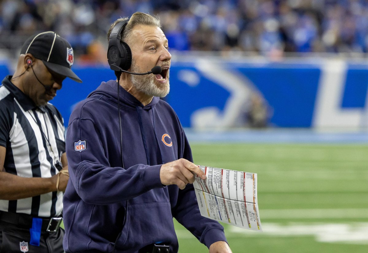 Ein Footballtrainer, der Matt Eberflus von den Bears sehr ähnlich sieht, trägt einen blauen Kapuzenpulli und ein Headset und schreit leidenschaftlich, während er an der Seitenlinie während einer gefühlten Thanksgiving-Niederlage eine Spieltabelle in der Hand hält. Ein Schiedsrichter in einem gestreiften Hemd steht daneben, während das Feld grün und in Vorfreude getaucht bleibt. Diese Beschreibung wurde mit der FootballR KI automatisch generiert.