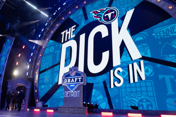 Cam Ward - NFL Draft Reihenfolge: Auf einer großen Bühne wird „The Pick Is In“ neben einem NFL Draft 2024-Logo gezeigt, wobei „Detroit“ prominent dargestellt ist. Im Hintergrund sind Tennessee Titans-Logos auf einer blauen Leinwand zu sehen, die auf die Reihenfolge des Drafts hinweisen. Helle Bühnenlichter beleuchten Menschen, die links neben einem kleinen Podium stehen. Diese Beschreibung wurde mit der FootballR KI automatisch generiert.