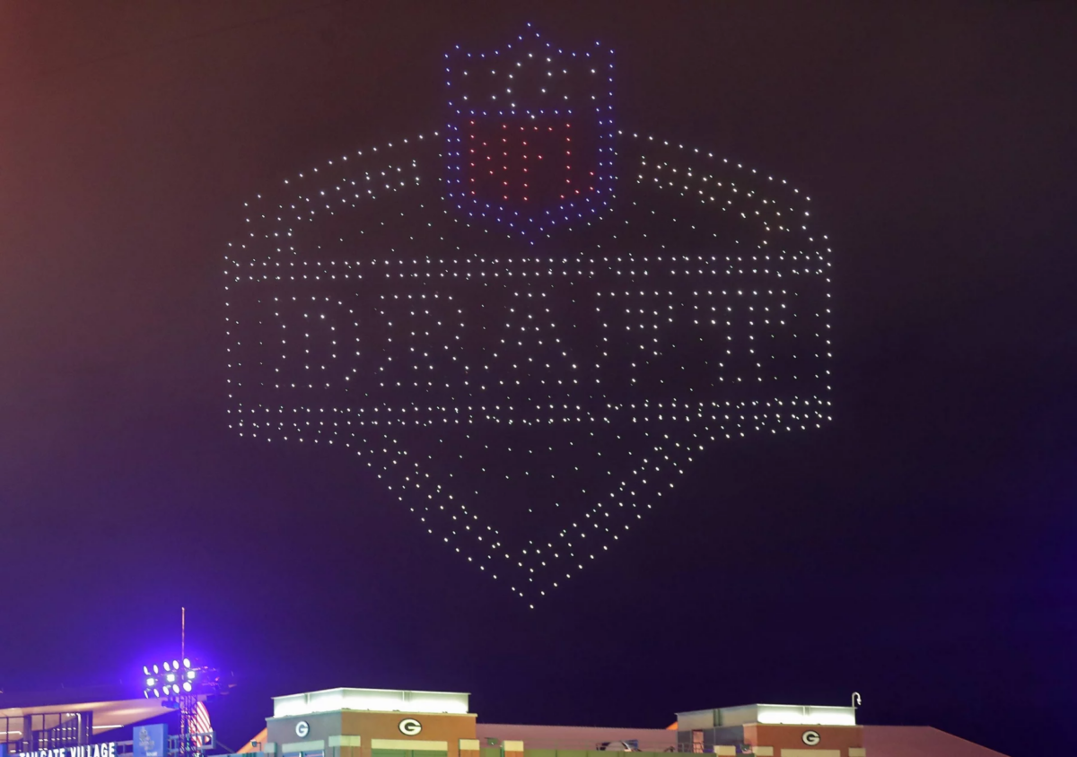 Derrick Harmon - Ein Nachthimmel wird von Hunderten Drohnen erleuchtet, die das NFL Draft-Logo bilden. „DRAFT“, das NFL-Schild und Derrick Harmons Name sind in Lichtern über grün dekorierten Gebäuden und dem „G“-Logo der Packers zu sehen. Diese Beschreibung wurde mit der FootballR KI automatisch generiert.