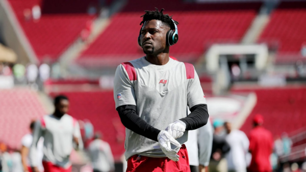 Ein Footballspieler, Antonio Brown, trägt ein graues Trikot, rote Shorts, weiße Handschuhe und große Kopfhörer trägt, steht auf einem fast leeren Feld. Konzentriert blickt er zur Seite, während sich im Hintergrund andere Spieler aufwärmen. Diese Beschreibung wurde mit der FootballR KI automatisch generiert.