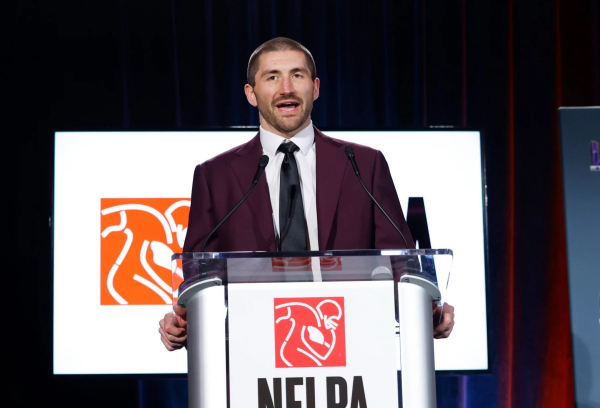 JC Tretter, in einem kastanienbraunen Anzug und mit schwarzer Krawatte, spricht an einem durchsichtigen Podium mit einem NFLPA-Logo. Hinter ihm zeigt ein Bildschirm dasselbe Logo mit einem stilisierten Football-Spieler in Weiß und Orange vor dunkelroter und blauer Hintergrundbeleuchtung. Diese Beschreibung wurde mit der FootballR KI automatisch generiert.