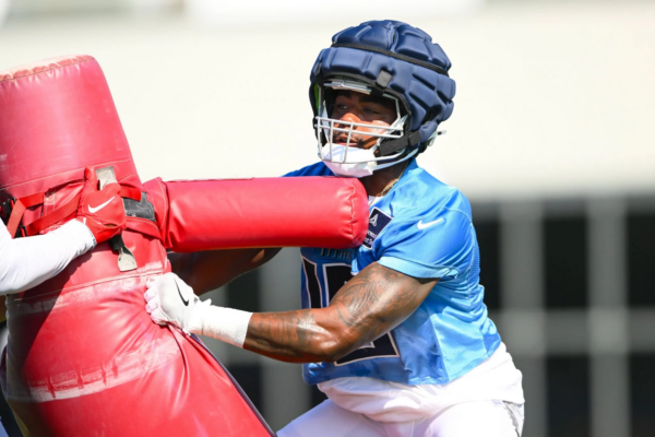 Camp - Treylon Burks, der ein blaues Trikot und einen gepolsterten Helm trägt, schiebt beim Training einen roten Tackling-Dummy. Er trägt weiße Handschuhe und sein Blick ist konzentriert. Der Hintergrund ist unscharf, um die Action und Intensität der Übung zu unterstreichen. Diese Beschreibung wurde mit der FootballR KI automatisch generiert.