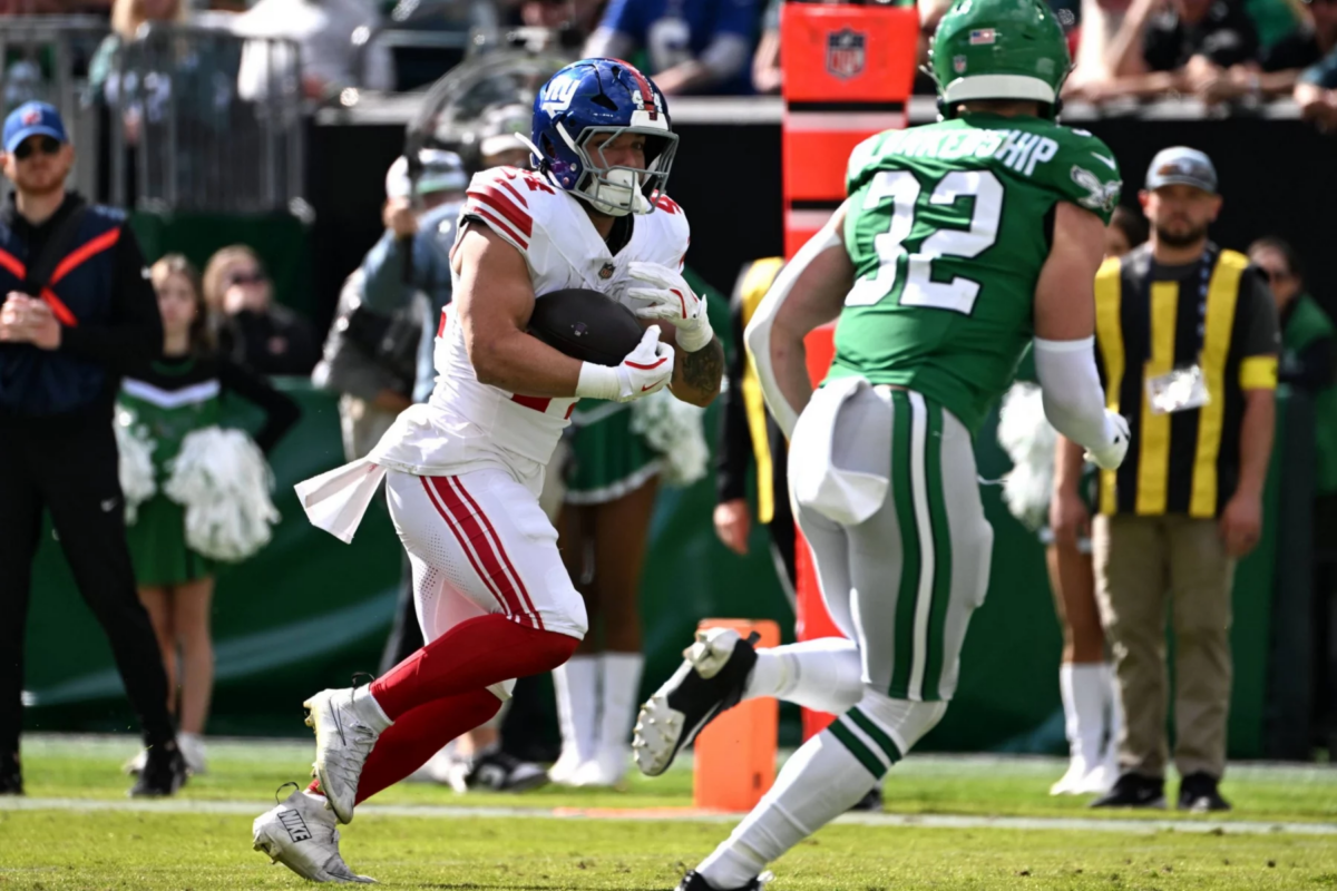 Cam Skattebo, New York Giants, in einer weißen Uniform läuft mit dem Ball, verfolgt von einem Spieler der Jets in Grün auf einem sonnigen Feld. Cheerleader und Offizielle sind zu sehen, während die Zuschauer von der Tribüne aus den Spielzug der Giants verfolgen. Diese Beschreibung wurde mit der FootballR KI automatisch generiert.