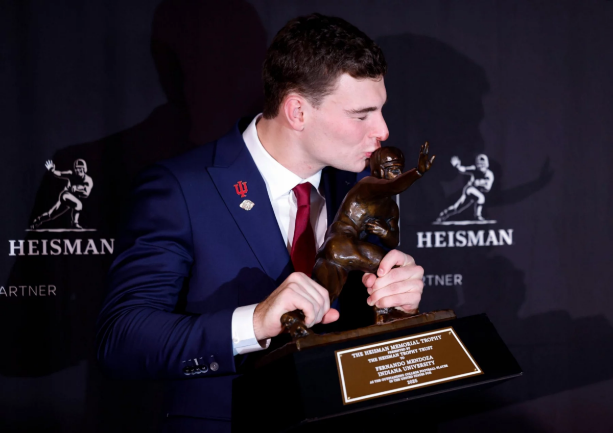 Fernando Mendoza, der eine rote Krawatte und eine Anstecknadel der Indiana University trägt, küsst die Heisman Trophy 2025 und hält sie mit beiden Händen. Im Hintergrund sind wiederholte Heisman-Logos zu sehen, und die Plakette der Trophäe ist deutlich zu erkennen. Diese Beschreibung wurde mit der FootballR KI automatisch generiert.