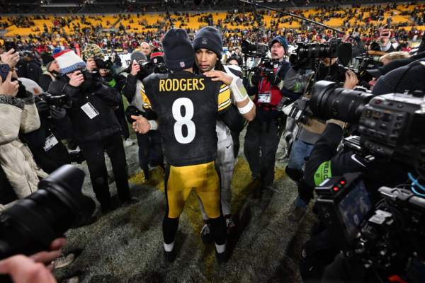 Houston Texans - Aaron Rodgers - Zwei American-Football-Spieler umarmen sich nach einem Spiel der Wild Card Round auf dem Spielfeld. Der Spieler, der der Kamera zugewandt ist, trägt ein schwarz-gelbes Steelers-Trikot mit der Aufschrift "RODGERS 8" auf dem Rücken, umgeben von Fotografen, Reportern und jubelnden Fans auf den Sitzen im Stadion. Diese Beschreibung wurde mit der FootballR KI automatisch generiert.