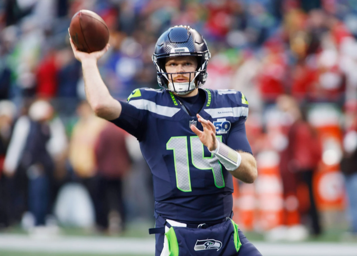 Sam Darnold - Ein Footballspieler im Trikot der Seattle Seahawks mit der Nummer 14 befindet sich mitten im Wurf während eines Spiels. Mit der Energie eines Playoff-Sieges trägt er einen dunklen Helm und hält den Football in der rechten Hand, während rot gekleidete Spieler im Hintergrund verschwimmen. Diese Beschreibung wurde mit der FootballR KI automatisch generiert.