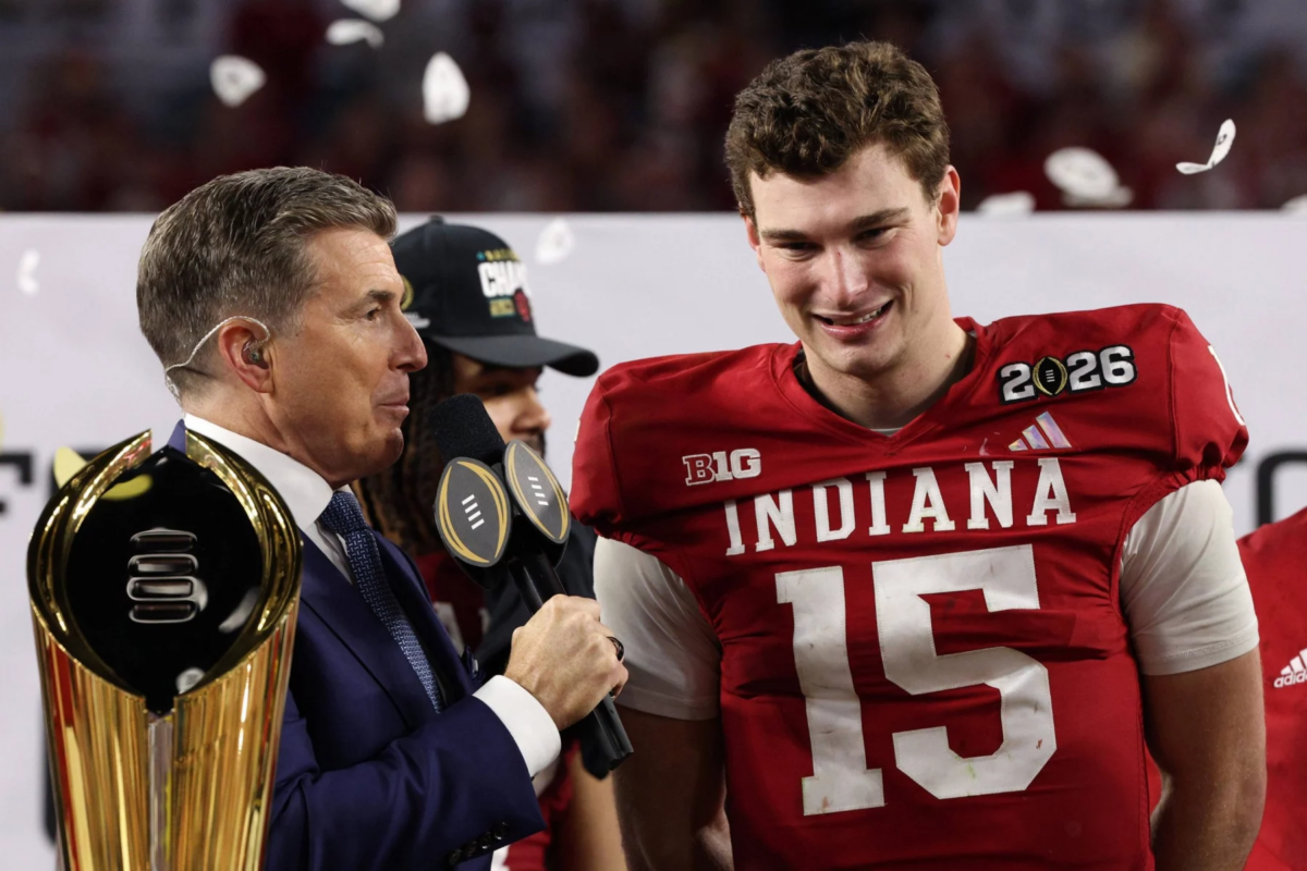 Ein lächelnder Indiana Footballspieler, Fernando Mendoza, mit der Trikotnummer 15 steht neben einem Interviewer, der ein Mikrofon hält. Konfetti fällt, während sie neben einer goldenen Meisterschaftstrophäe den nationalen Titel feiern. Der Spieler trägt einen Aufnäher mit der Aufschrift "2026"; das Stadion hinter ihnen ist unscharf. Diese Beschreibung wurde mit der FootballR KI automatisch generiert.