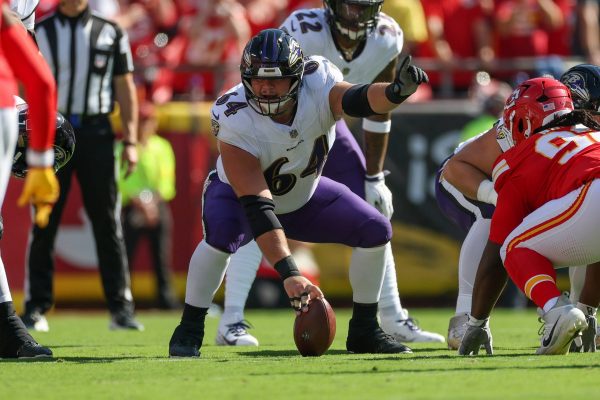 Tyler Linderbaum - Ein Football-Spieler der Offensive Line der Baltimore Ravens in einer weiß-lila Uniform bereitet sich während eines Spiels gegen die Kansas City Chiefs darauf vor, den Ball an der Line of Scrimmage zu spielen. Alt-Text wurde KI-generiert.