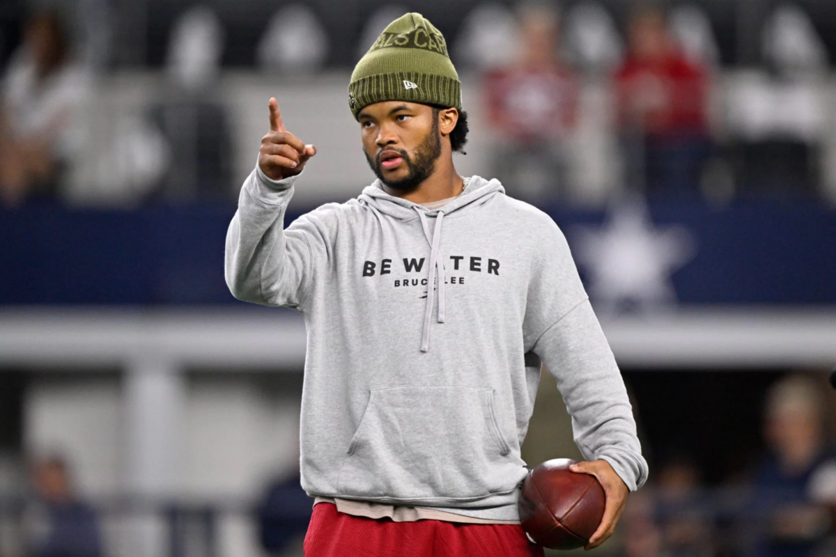 Kyler Murray, der einen grauen "Be Water Bruce Lee"-Kapuzenpulli und eine olivfarbene Mütze trägt, hält auf dem Football-Feld der Cardinals einen Football in der Hand und zeigt nach vorne, um die Energie von Kyler Murray zu kanalisieren. Alt-Text wurde KI-generiert.