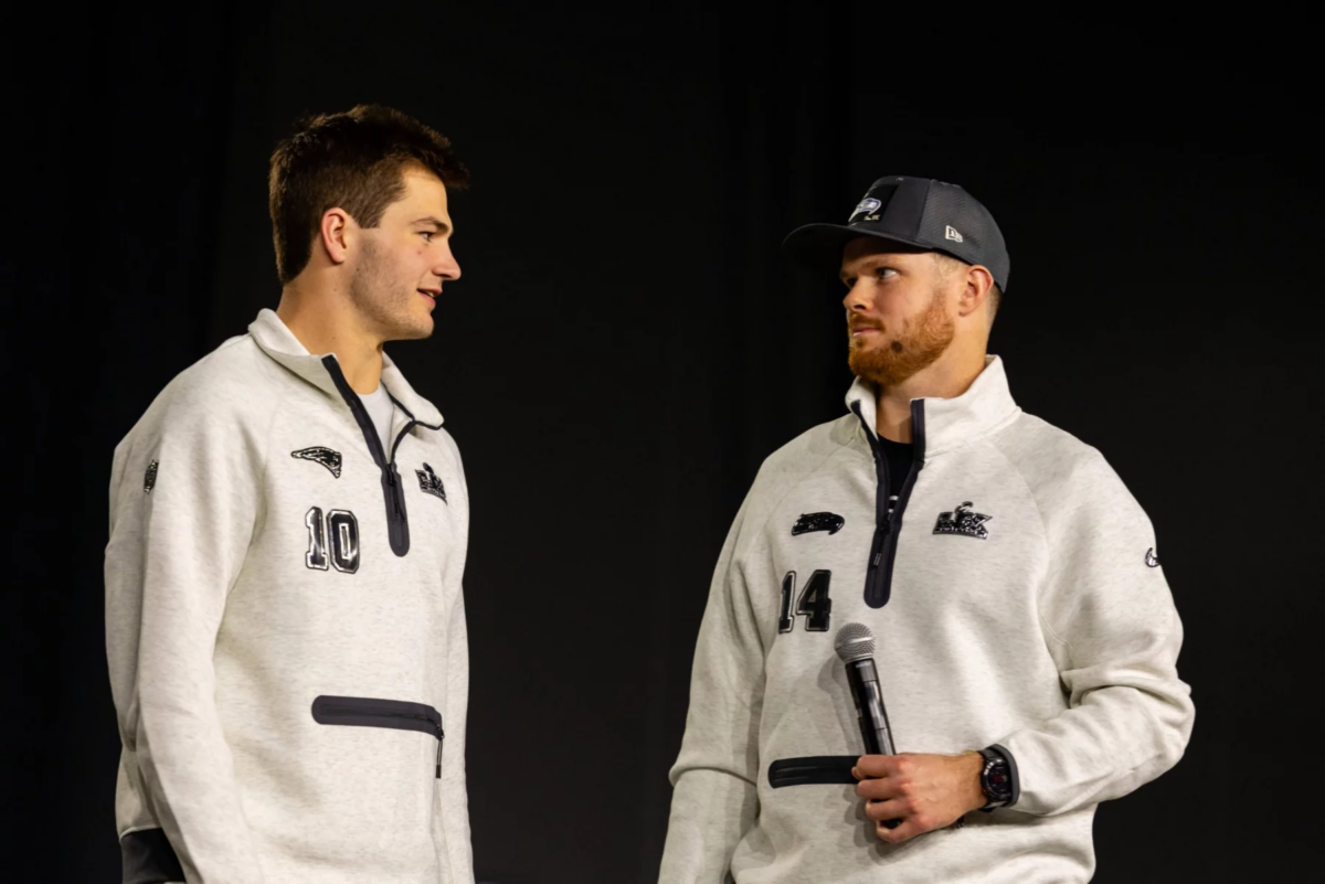 Drake Maye und Sam Darnold - Zwei Männer in passenden hellgrauen Pullovern, von denen einer ein Mikrofon in der Hand hält, stehen sich vor einem schlichten dunklen Hintergrund gegenüber und fangen die Aufregung der Eröffnungsfeier des Super Bowl LX ein. Alt-Text wurde KI-generiert.