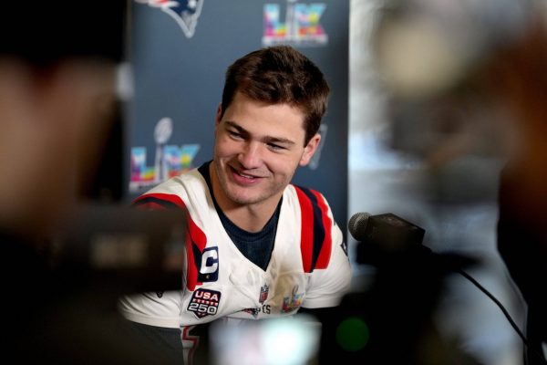 Drake Maye - Ein Footballspieler im Trikot der New England Patriots spricht auf einer Pressekonferenz, lächelnd, mit Mikrofonen und Kameras vor sich, über die Super Bowl-Ambitionen des Teams. Alt-Text wurde KI-generiert.