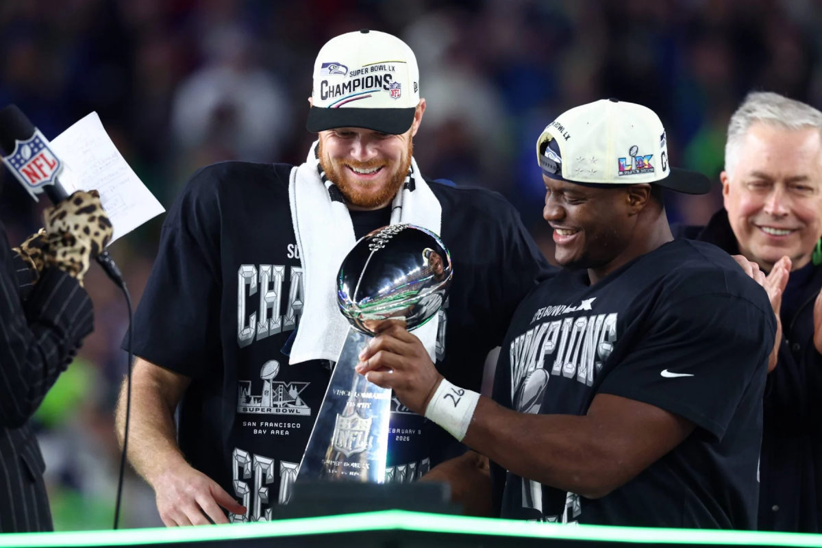 Sam Darnold und Kenneth Walker III - Zwei Football-Spieler mit "Champions"-Hüten und -Shirts lächeln, als sie während der Super Bowl 50-Feier die Vince Lombardi Trophy halten und damit beweisen, dass alles möglich ist, wenn du an dich selbst glaubst. Alt-Text wurde KI-generiert.