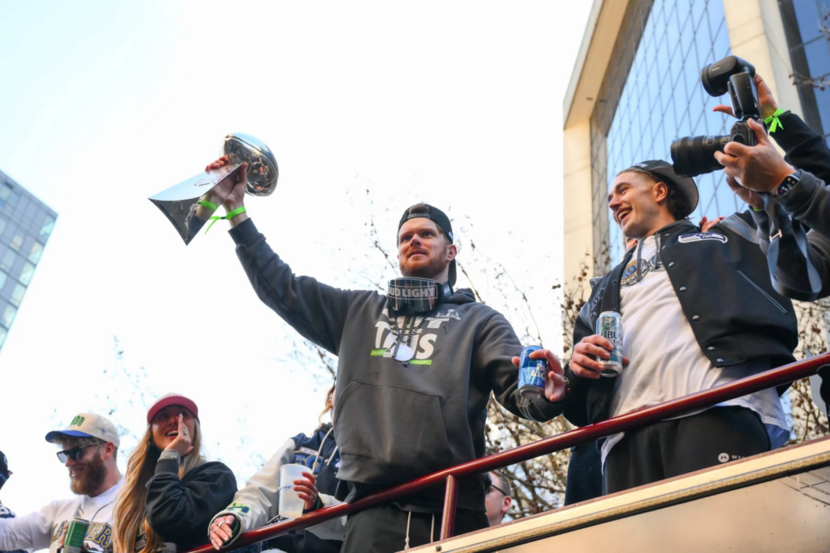 Sam Darnold hält eine Trophäe auf einem Umzugswagen in Seattle hoch, umgeben von anderen Personen, von denen einige Fotos machen und Getränke in der Hand halten, um den Super-Bowl-Sieg der Seahawks zu feiern. Alt-Text wurde KI-generiert.
