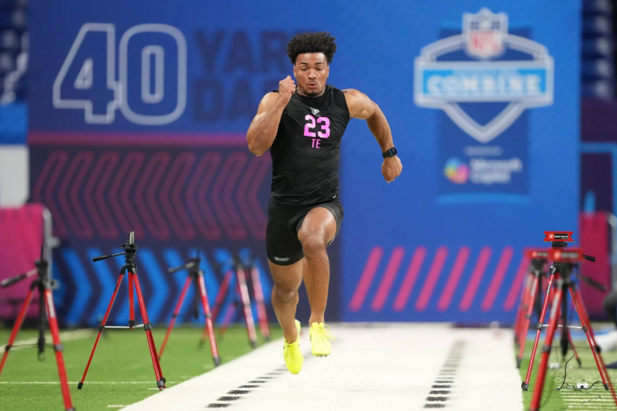 Kenyon Sadiq - Ein Athlet in einem schwarzen Outfit mit der Aufschrift "23 TE" absolviert beim NFL Scouting Combine 2026 einen 40-Yard-Lauf, umgeben von Zeitmessgeräten auf einem Hallenfeld neben anderen herausragenden Tight Ends und Defensive Backs. Alt-Text wurde KI-generiert.
