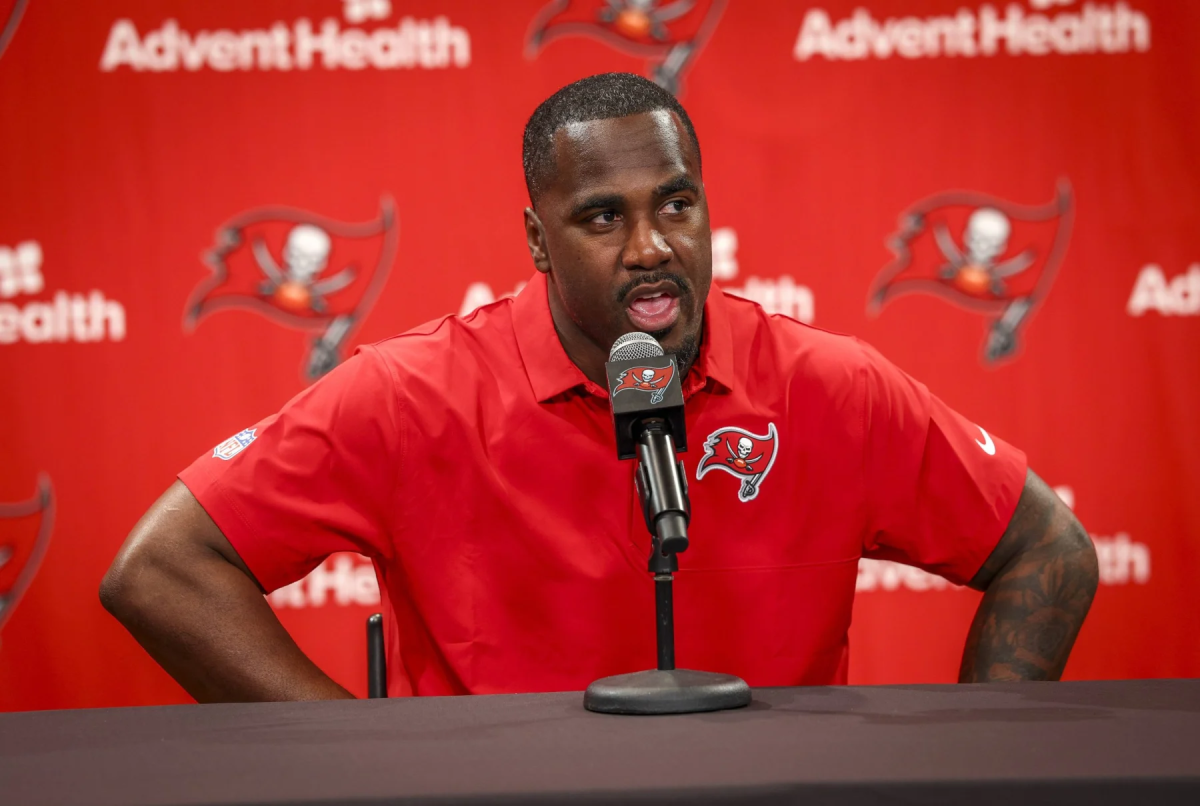 Lavonte David in rotem Tampa Bay Buccaneers Poloshirt spricht bei Pressekonferenz vor rotem AdventHealth-Hintergrund