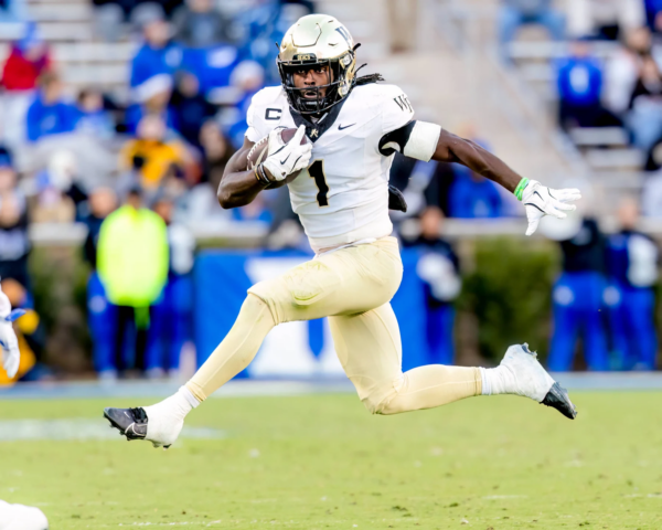 NFL Draft 2026 Kandidat Demond Claiborne zeigt seine Fähigkeiten als Footballspieler in einem weiß-goldenen Trikot, indem er in der Luft springt, während er mit dem Ball läuft - ein beeindruckender Moment für sein Spielerprofil. Alt-Text wurde KI-generiert.