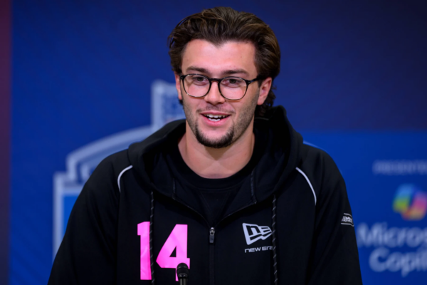 Ein Mann mit Brille und dunklem Haar, der einen schwarzen Kapuzenpulli mit der Nummer 14 in Rosa trägt, spricht während des NFL Draft 2026 in ein Mikrofon vor blauem Hintergrund. Alt-Text wurde KI-generiert.