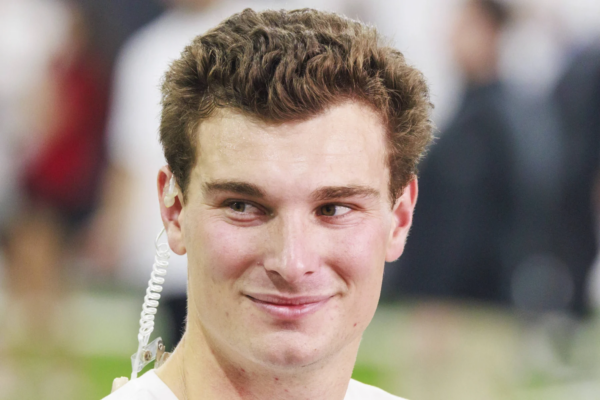 Ein junger Mann mit kurzen braunen Haaren, der ein weißes Hemd und einen Ohrhörer trägt, schaut leicht zur Seite und lächelt. Der Hintergrund ist unscharf und deutet auf Fernando Mendozas Begeisterung für den NFL Draft 2026 und seine Träume mit den Raiders hin. Alt-Text wurde KI-generiert.