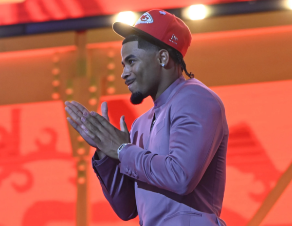 Mansoor Delane, ein Cornerback, faltet seine Hände zusammen, während er eine rote Kansas City Chiefs-Mütze und einen lilafarbenen Anzug trägt und vor einem leuchtend roten und orangen Hintergrund steht. Alt-Text wurde KI-generiert.