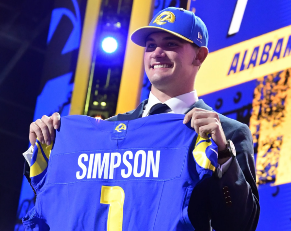 Ein lächelnder Mann im Anzug und mit Teamkappe hält ein blaues Rams-Football-Trikot mit der Aufschrift "Simpson" und der Nummer 1 hoch und feiert damit Ty Simpson als potenziellen Stafford-Nachfolger bei einer Draft-Veranstaltung. Alt-Text wurde KI-generiert.