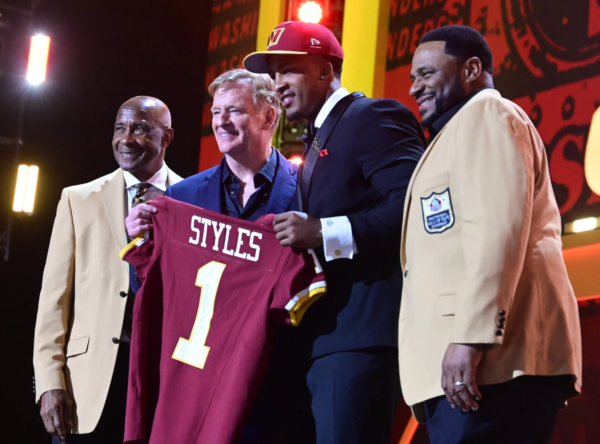 Vier Männer stehen auf der Bühne, einer von ihnen hält ein kastanienbraunes Football-Trikot mit der Aufschrift "STYLES 1". Zwei Männer tragen hellbraune Jacken mit Hall of Fame-Aufnähern, während der NFL Draft 2026 die Big Ten-Aussichten der Rams hervorhebt. Alt-Text wurde KI-generiert.