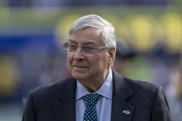 Ein älterer Mann mit grauen Haaren und Brille trägt einen Anzug, ein blaues Hemd und eine Krawatte mit Polka-Dots. Er steht im Freien vor einem unscharfen Hintergrund und strahlt dabei einen dezenten Stolz auf die Buffalo Bills aus. Alt-Text wurde KI-generiert.