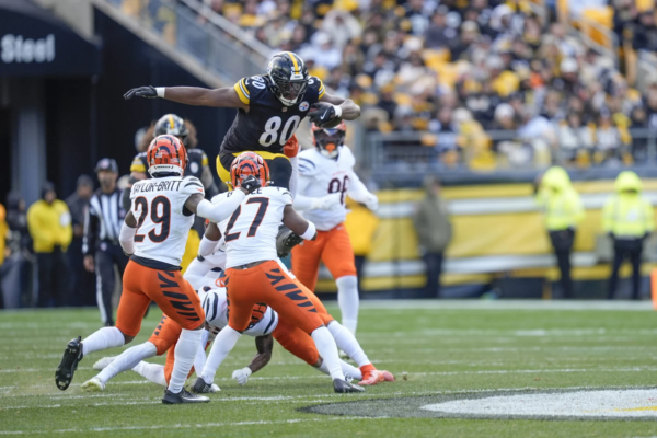 Ein Spieler der Pittsburgh Steelers, inspiriert von Darnell Washington kürzlicher Vertragsverlängerung, springt während eines Footballspiels an einem sonnigen Tag über zwei Verteidiger der Cincinnati Bengals. Alt-Text wurde KI-generiert.