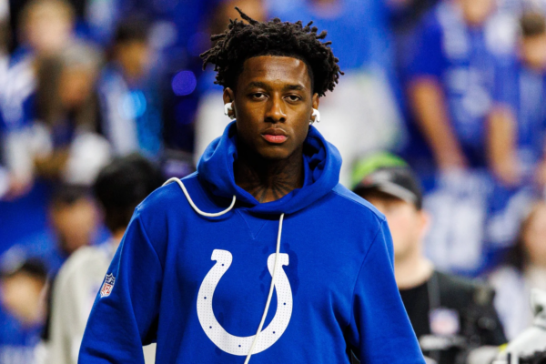 Ein Mann, der einen blauen Indianapolis-Colts-Hoodie und Kopfhörer trägt, steht in einem Stadion, im Hintergrund sind verschwommene Zuschauer zu sehen; möglicherweise streamt er inmitten der Menge Internet-Highlights von Sauce Gardner. Alt-Text wurde KI-generiert.