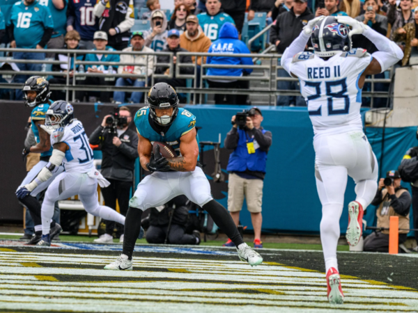 Brenton Strange, Tight End der Jaguars, im blaugrünen Trikot, fängt einen Pass in der Endzone, während ein Verteidiger in Weiß die Arme hebt; Zuschauer und Fotografen beobachten das Geschehen von der Seitenlinie aus. Alt-Text wurde KI-generiert.