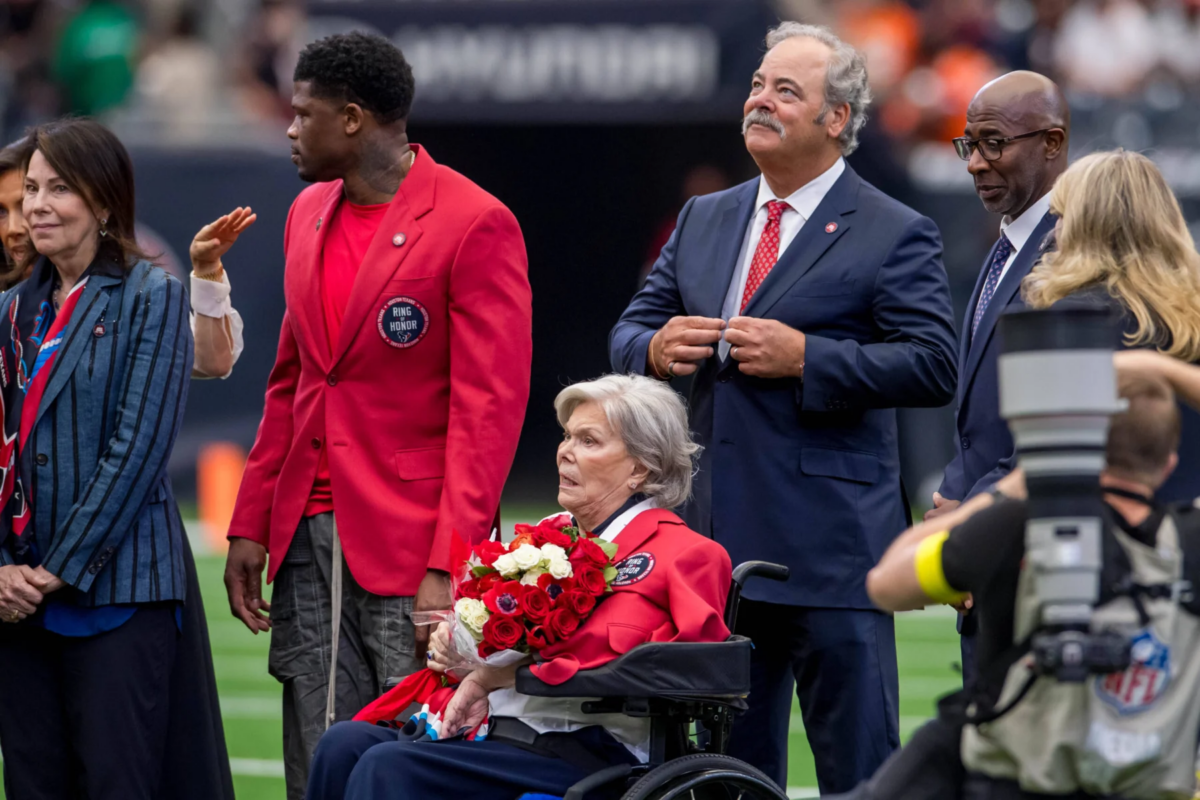 Eine Frau im Rollstuhl, die Rosen in den Händen hält und als Janice McNair identifiziert wurde, ist auf dem Footballfeld während einer Feier der Houston Texans zu sehen, umgeben von Menschen in Anzügen und Jacken. Alt-Text wurde KI-generiert.