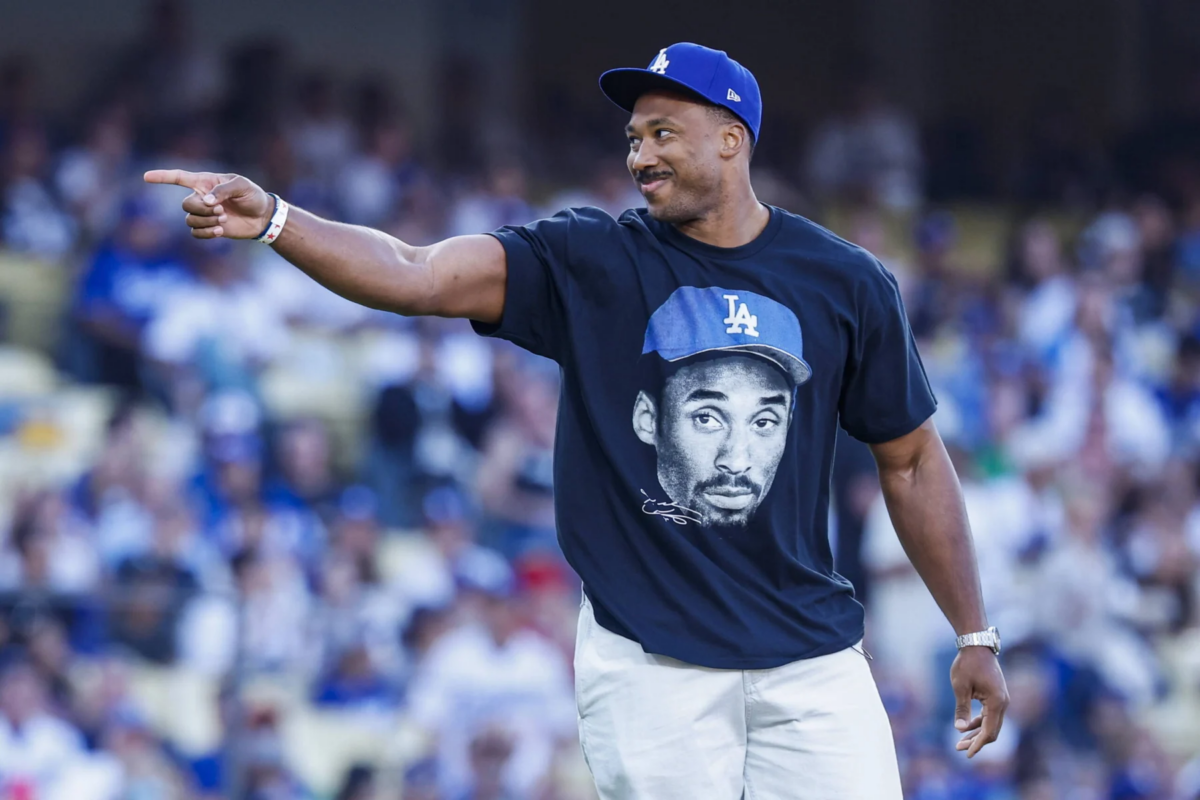 Ein Mann mit einer LA-Dodgers-Kappe und einem T-Shirt mit einem aufgedruckten Gesicht, der auf einem Baseballfeld nach links zeigt, im Hintergrund die Zuschauermenge – ein Bild, das die Aufregung eines Spieltags einfängt und an Rams-Fans erinnert, die Myles Garrett bei den „Camp-Tag“-Veranstaltungen entdecken. Alt-Text wurde KI-generiert.