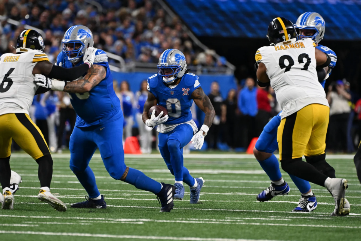Jahmyr Gibbs, ein Runningback der Detroit Lions, läuft mit dem Football, während seine Teamkollegen und die Verteidiger der Pittsburgh Steelers während eines NFL-Spiels in einen Zweikampf verwickelt sind. Alt-Text wurde KI-generiert.