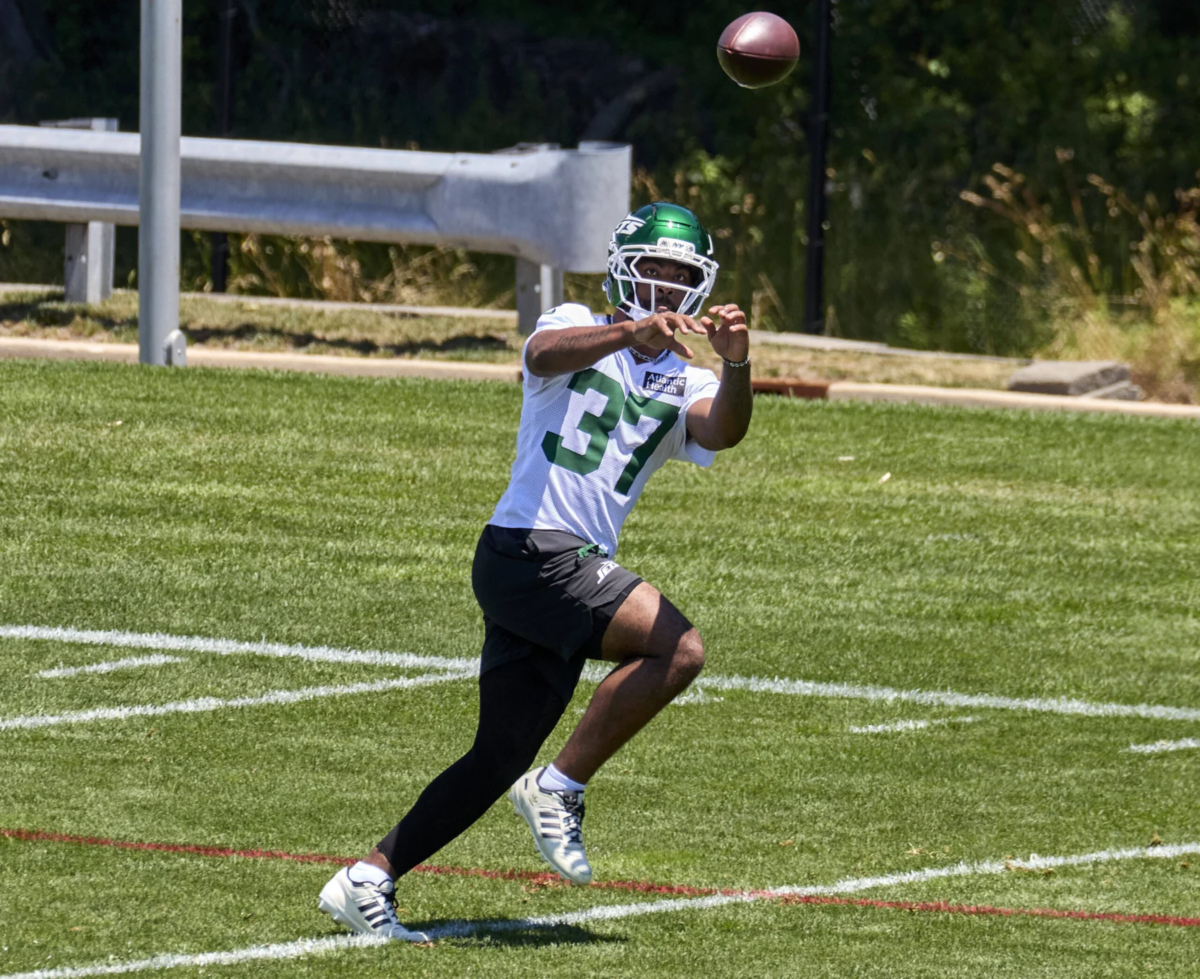 Ein Footballspieler mit weißem Helm und grünem Jets-Trikot mit der Nummer 37, Qwan’tez Stiggers, rennt während des Trainings über das Spielfeld und fängt dabei einen Football. Alt-Text wurde KI-generiert.