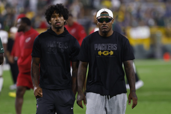 Zwei Männer in Packers-Trikots stehen nebeneinander auf einem Footballfeld, Josh Jacobs trägt eine Sonnenbrille und eine Kappe; im Hintergrund sind verschwommene Menschen und Stadionlichter zu sehen, während LaFleur erwartet, dass die Packers eine starke Saison spielen werden. Alt-Text wurde KI-generiert.