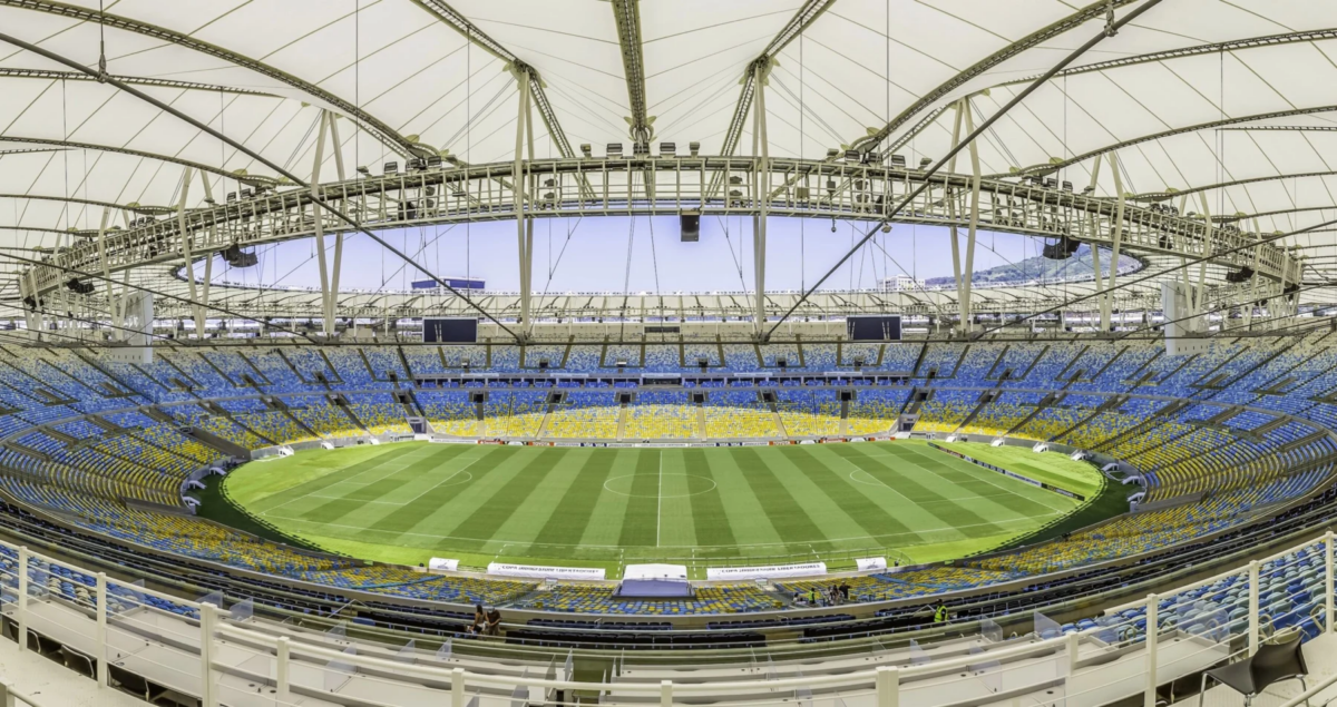 Rio de Janeiro- Weitwinkelaufnahme eines leeren, modernen Stadions mit einem teilweise überdachten Dach, einem grünen Rasenfeld und blauen und gelben Sitzplätzen rund um das Spielfeld; die NFL ist zuversichtlich, dass die Platzverhältnisse für die bevorstehenden Veranstaltungen optimal sind. Alt-Text wurde KI-generiert.