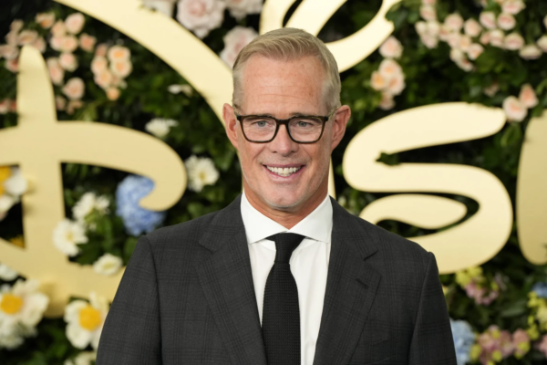 Joe Buck - Ein Mann in einem dunklen Anzug, einem weißen Hemd und einer schwarzen Krawatte steht vor einem blumigen Hintergrund mit goldener Schrift und lächelt in die Kamera – eine Szene, die an eine feierliche Veranstaltung erinnert, bei der Ankündigungen im Stil von „Bericht: Joe Buck einigt sich“ gemacht werden könnten. Alt-Text wurde KI-generiert.