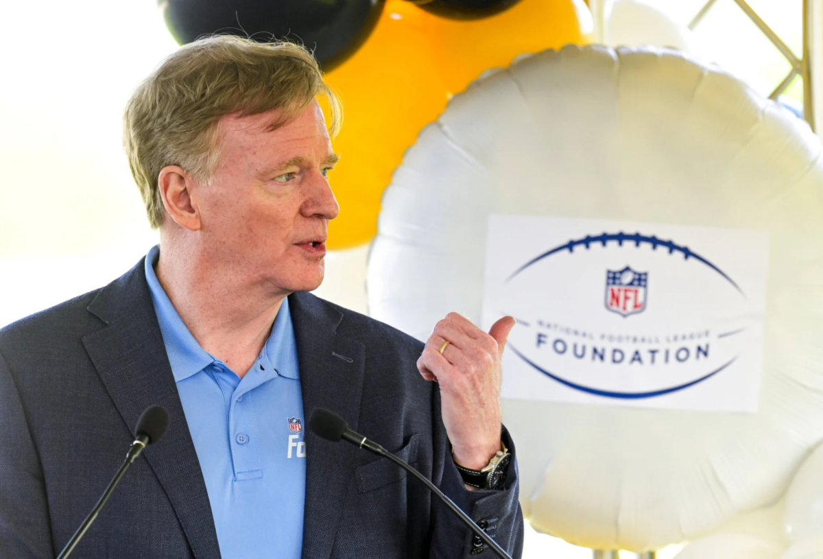Roger Goodell - Ein Mann in einem blauen Hemd und einem dunklen Blazer spricht ins Mikrofon und gestikuliert dabei mit der Hand; hinter ihm ist ein Luftballon mit dem Logo der NFL Foundation zu sehen, der an die jüngsten Ereignisse erinnert, bei denen Goodell eine Vertragsverlängerung unterzeichnet hat. Alt-Text wurde KI-generiert.