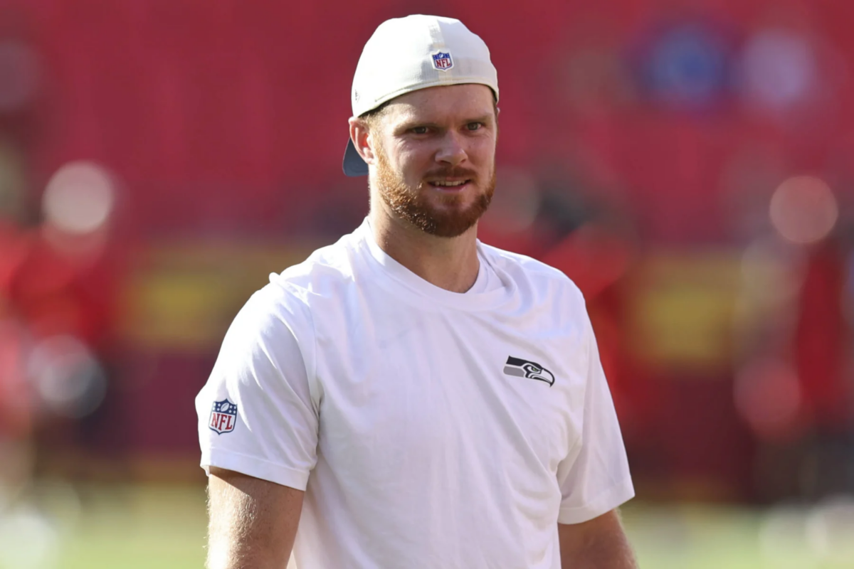 Sam Darnold - Ein Mann in einem weißen Seahawks-Trikot und mit einer nach hinten gedrehten Kappe steht auf einem Footballfeld vor einem verschwommenen roten Hintergrund und bereitet sich möglicherweise darauf vor, nach dem Spiel einen Bericht über die Seahawks zu liefern. Alt-Text wurde KI-generiert.