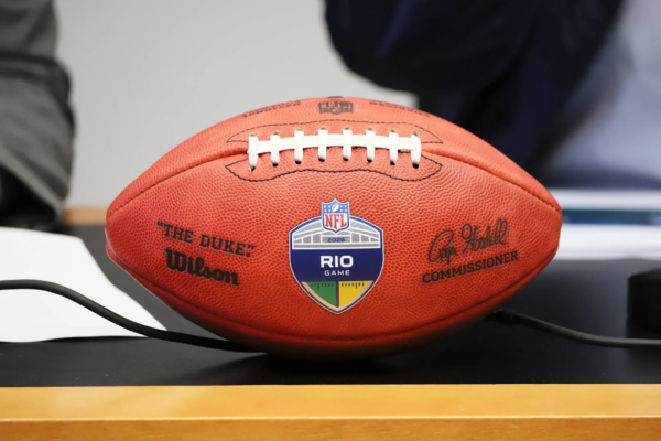 Rio de Janeiro - Ein Wilson-NFL-Football mit der Aufschrift „The Duke“ und einem „RIO GAME“-Aufkleber liegt auf einem Tisch; auf dem Ball sind Unterschriften und Logos zu sehen – Kein Grund zur Sorge, dieses wertvolle Sammlerstück ist gut geschützt und bereit für die Ausstellung. Alt-Text wurde KI-generiert.