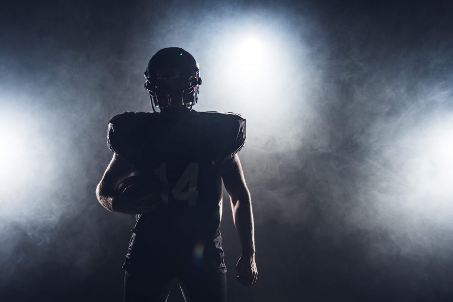 FootballR - NFL - Die Silhouette eines Footballspielers, der einen Ball hält, vor einem dunklen Hintergrund, perfekt, um Ihr DAZN-Abo zu präsentieren.