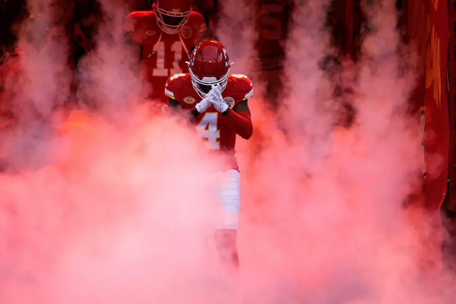 FootballR - NFL Rashee Rice - Ein Wide Receiver der Kansas City Chiefs in einer roten Uniform mit der Nummer 14 taucht dramatisch aus einer roten Rauchwolke auf, im Hintergrund sind teilweise Teamkollegen zu sehen, was einen intensiven Moment vor dem Spiel auf dem Diese Beschreibung wurde automatisch generiert.