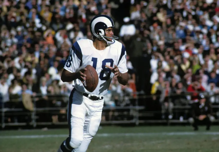 FootballR - NFL - Ein Quarterback in weiß-blauer Uniform mit der Nummer 18, identifiziert als NFL MVP Roman Gabriel, sucht während eines sonnigen Footballspiels nach einem freien Receiver. Er hält den Football in seiner rechten Diese Beschreibung wurde automatisch generiert.
