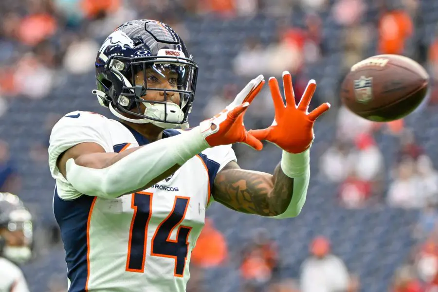 FootballR - NFL - Courtland Sutton, ein Spieler der Denver Broncos mit der Nummer 14, streckt während eines Spiels die Hand aus, um einen Football zu fangen. Er ist hochkonzentriert und seine orangefarbenen Handschuhe stechen von seiner blau-weißen Uniform hervor. Diese Beschreibung wurde automatisch generiert.