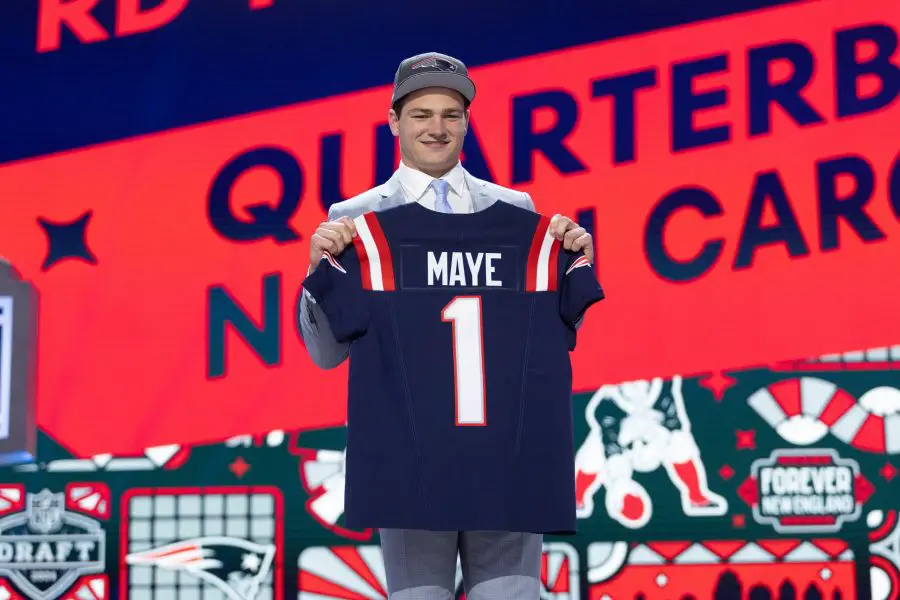 FootballR - NFL - Ein junger Mann auf einer Bühne beim NFL Draft 2024, lächelt und hält ein Footballtrikot mit Drake Maye und der Nummer „1“ hoch. Ein leuchtend roter und blauer Hintergrund Diese Beschreibung wurde automatisch generiert.