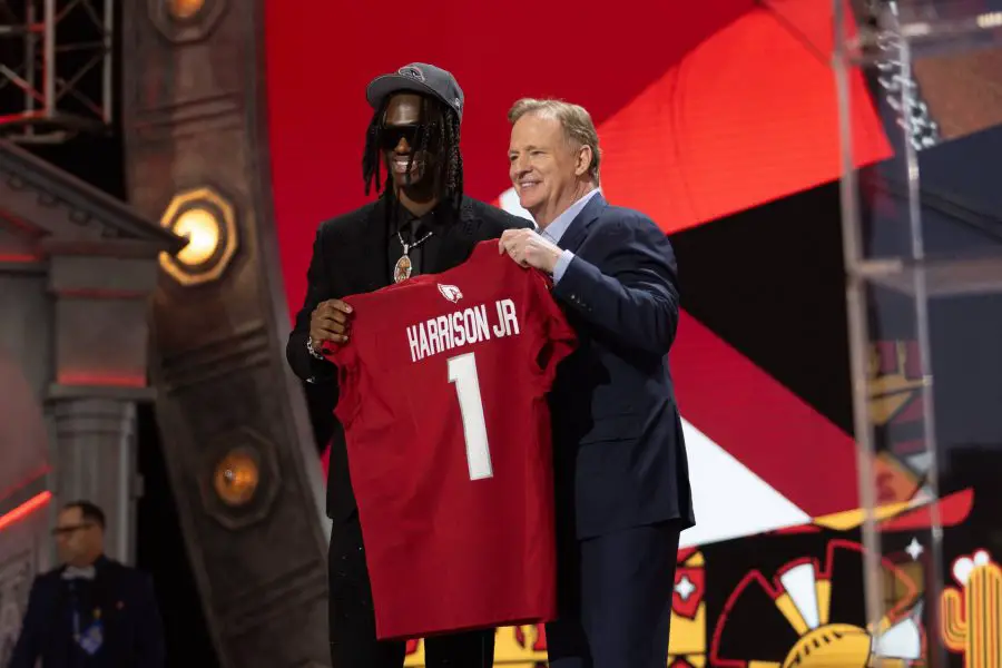 FootballR - NFL - Zwei Männer auf einer Bühne, einer jung in einer bestickten Jacke und Mütze, lächelnd, während er ein rotes Footballtrikot mit der Aufschrift Marvin Harrison Jr. 1 aus dem NFL Draft 2024 hält. Diese Beschreibung wurde automatisch generiert.