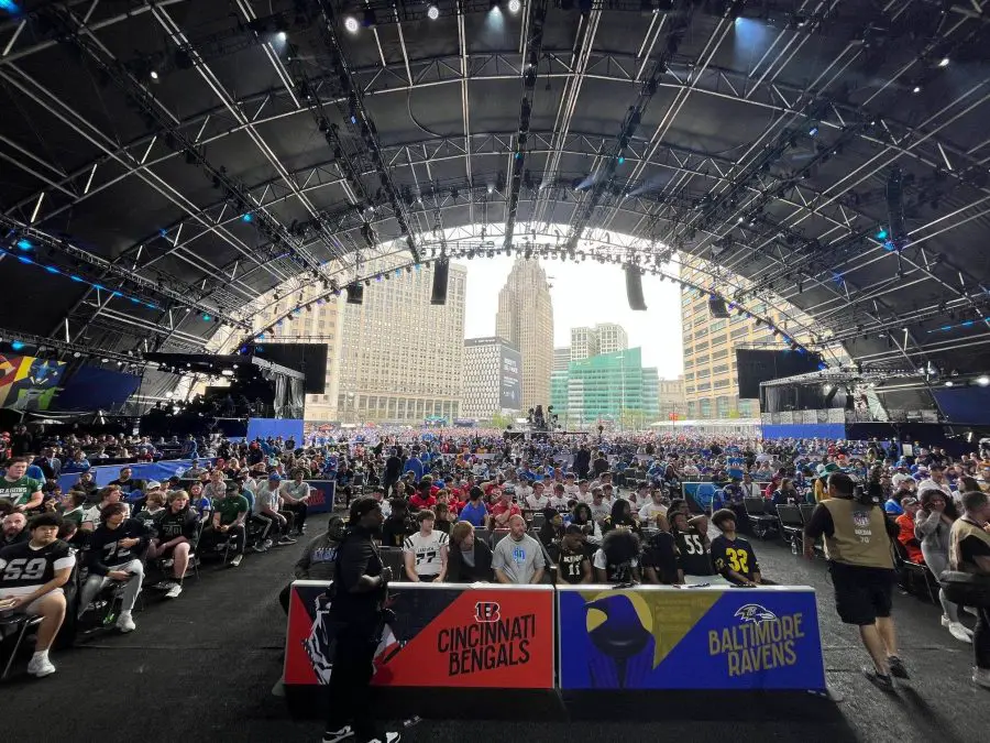 FootballR - NFL - Detroit - Eine große Menschenmenge versammelte sich in einem weitläufigen Veranstaltungsort mit hoher, gewölbter Decke für den NFL Draft 2024. Auf der Bühne hängen Banner der Cincinnati Bengals und Baltimore Ravens. Eine Stadtkulisse fügt Diese Beschreibung wurde automatisch generiert.